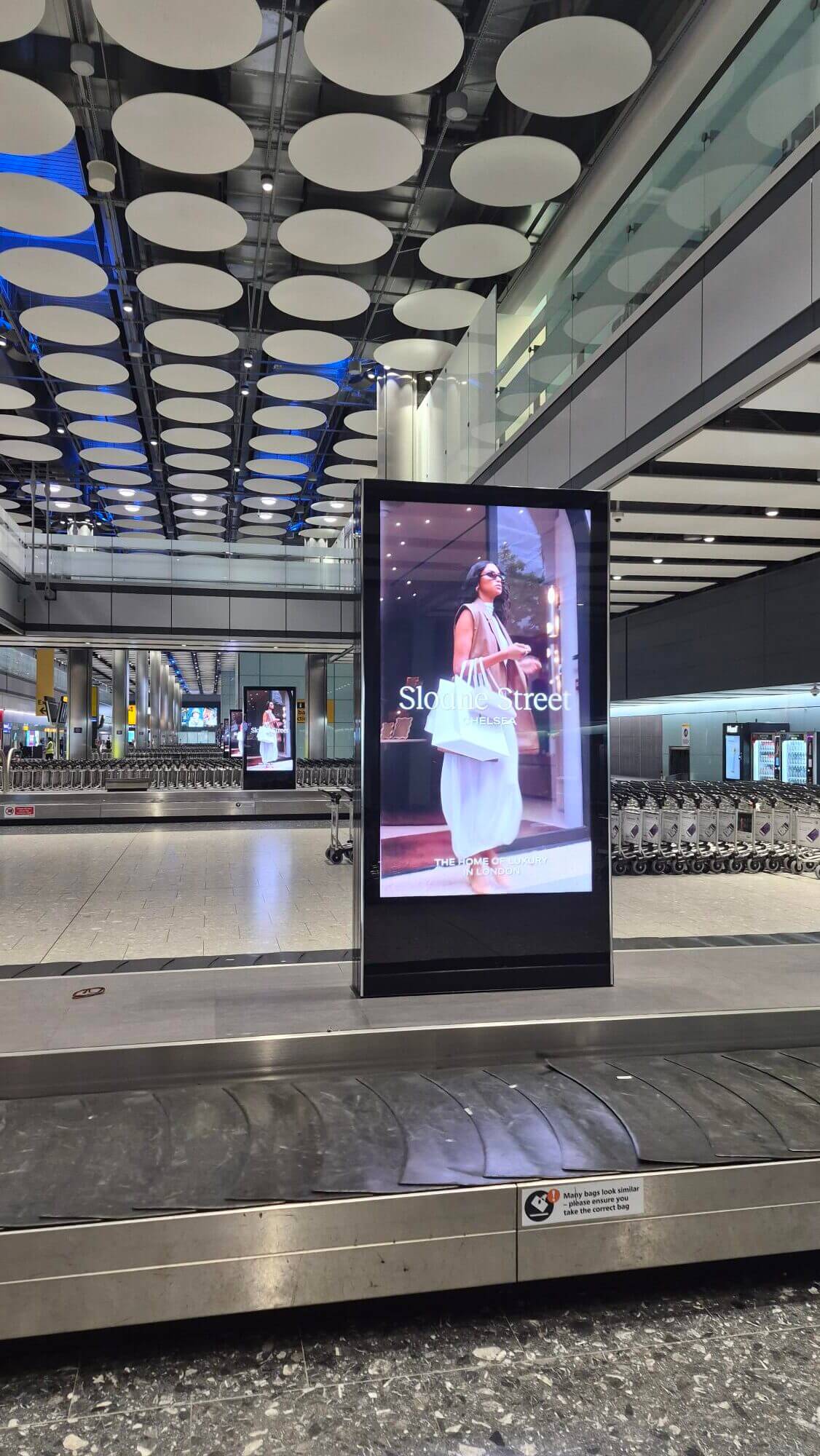Traveller carrying Sloane Street shopping bags, part of an airport OOH advertising creative on a digital totem unit at London Heathrow baggage reclaim